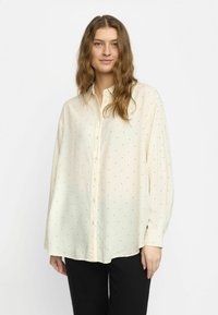 Hellbeige Hemd mit schwarzen Pünktchen, durchgehende Knopfleiste, lockere Passform, lange Ärmel und abgerundetem Saum. Der Stoff wirkt glatt und weich.