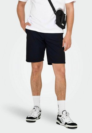 Schwarze Shorts aus fabric, mit geradem Schnitt und Seitentaschen. Getragen mit einem weißen T-Shirt, einer schwarzen Umhängetasche und Turnschuhen.
