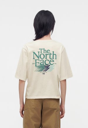 Persona con cabello oscuro lleva una camiseta beige con "The North Face" y un gráfico en la espalda, combinada con pantalones marrones, mirando de espaldas a la cámara.