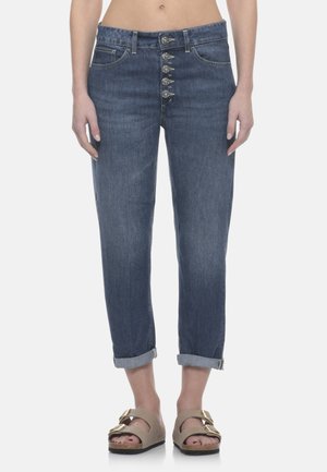 Donna con jeans blu risvoltati a cinque bottoni visibili, in piedi a piedi nudi con sandali beige con fibbia, ritratta dalla vita alle caviglie.