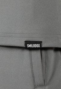 Tejido gris con una textura suave. Presenta una etiqueta de marca negra con la inscripción "SMILODOX" y un bolsillo oculto en el lado.