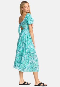 Une robe mi-longue en teal clair avec un motif floral, des manches bouffantes, un dos découpé et un détail noué, associée à des sandales noires.