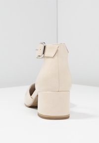 Béžová lodička s kulatou špičkou, hladkým povrchem a plochým blokovým podpatkem. Obsahuje pásek kolem kotníku se stříbrnou kovovou sponou.