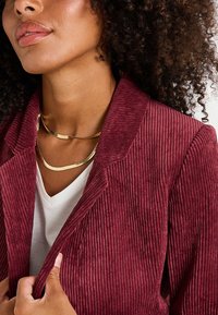 Blazer de pana burdeos con solapas anchas y estrías verticales, superpuesto a una camiseta blanca de escote en V, complementado con un collar dorado.