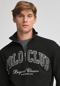 Svart zip-up sweatshirt med en stor "POLO CLUB" grafik i grå och vit text. Tyget verkar mjukt med en stående krage.