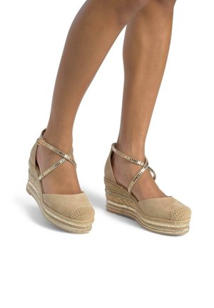 Beige espadrille wedge sandals with cross-straps and braided jute midsole. Suede upper with a textured toe cap and metallic accents.