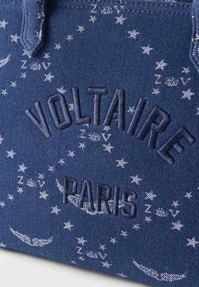 Μπλε ύφασμα με κεντημένο κείμενο «Voltaire Paris» περιτριγυρισμένο από λευκά μοτίβα αστέρι και φτερών σε κυκλικό σχέδιο.