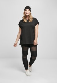 T-shirt noir oversized avec des manches roulées, associé à des leggings noirs transparents et côtelés, complété par un bonnet en tricot noir et des baskets blanches.