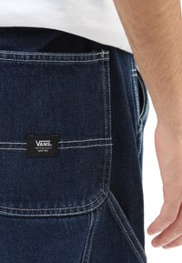 Pantalon en denim avec un lavage foncé, arborant une étiquette rectangulaire noire avec du texte blanc sur la poche arrière. Des surpiqûres contrastées sont visibles.