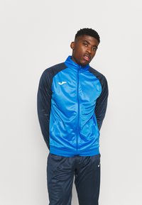 Giacca sportiva blu con zip, maniche nere a contrasto, texture liscia, con logo sul lato sinistro del petto. Abbinata a pantaloni coordinati.