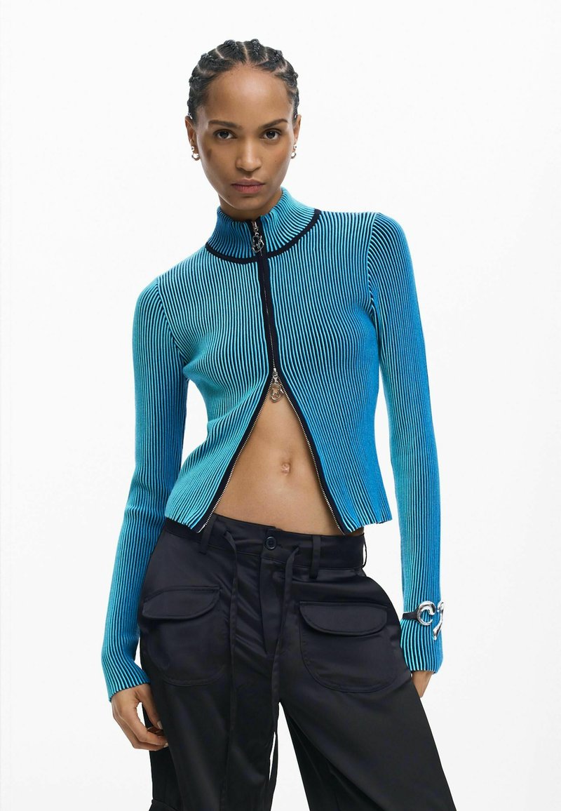 Niebieska żebrowana dzianinowa kurtka zapinana na zamek z wysokim kołnierzem i kontrastowymi czarnymi akcentami, zestawiona z czarnymi spodniami typu cargo z bocznymi kieszeniami.