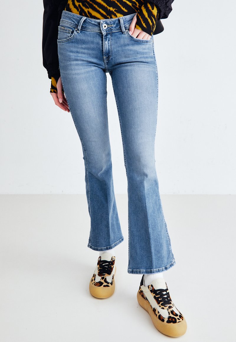 Jean flare bleu clair avec un délavage moyen, présentant une fermeture à bouton et zip, des poches frontales et un ourlet propre. Associé à des baskets à motifs.