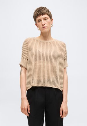 Kvinde med kort hår iført beige strikket kortærmet sweater og sorte bukser, stående mod en lys baggrund med neutralt ansigtsudtryk.