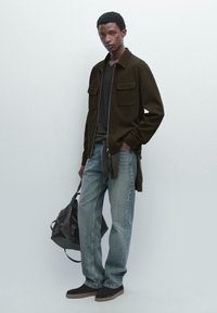 Mørkegrønt jakke med to brystlommer, grå skjorte, lyseblå jeans og mørke sko. Sort taske i hånden. Enkel og moderne design.