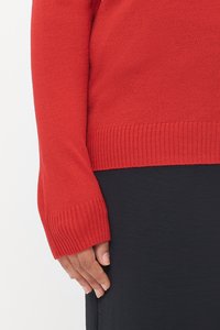 Pulôver de malha vermelho com mangas longas e punhos canelados, combinado com uma peça preta. Foco na textura e no contraste de cores.