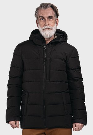 Älterer Mann mit grauem Haar und Bart, der eine schwarze gesteppte Winterjacke mit Kapuze trägt, stehend vor einem einfarbigen hellen Hintergrund.