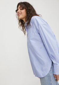 Camicia a righe blu e bianche con una vestibilità morbida, maniche lunghe e una tasca sul lato sinistro, abbinata a jeans in denim azzurro chiaro.