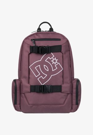 Mochila marrón con correas y hebillas negras. Presenta un gran logo blanco en la parte frontal. Hecha de un tejido duradero con bolsillos laterales con cremallera.