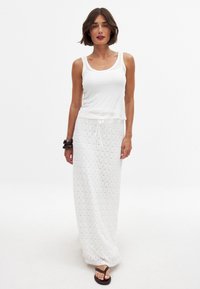 Un débardeur blanc à larges bretelles associé à une jupe blanche à motifs longueur cheville. Des sandales plates noires complètent le look.