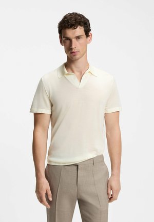 Man with short curly hair wears a light cream short-sleeve knit polo shirt and beige tailored trousers, standing against a plain background.