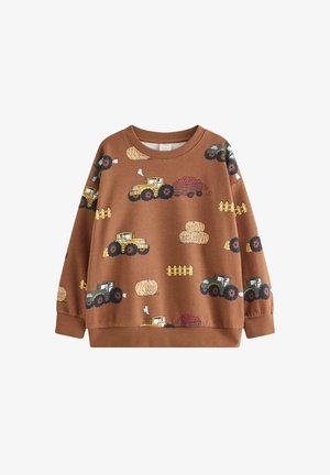 Lille barn med krøllet blondt hår iført en brun sweater med traktormønster, sorte bukser og brune sko med blå stropper, stående på en ensfarvet baggrund.