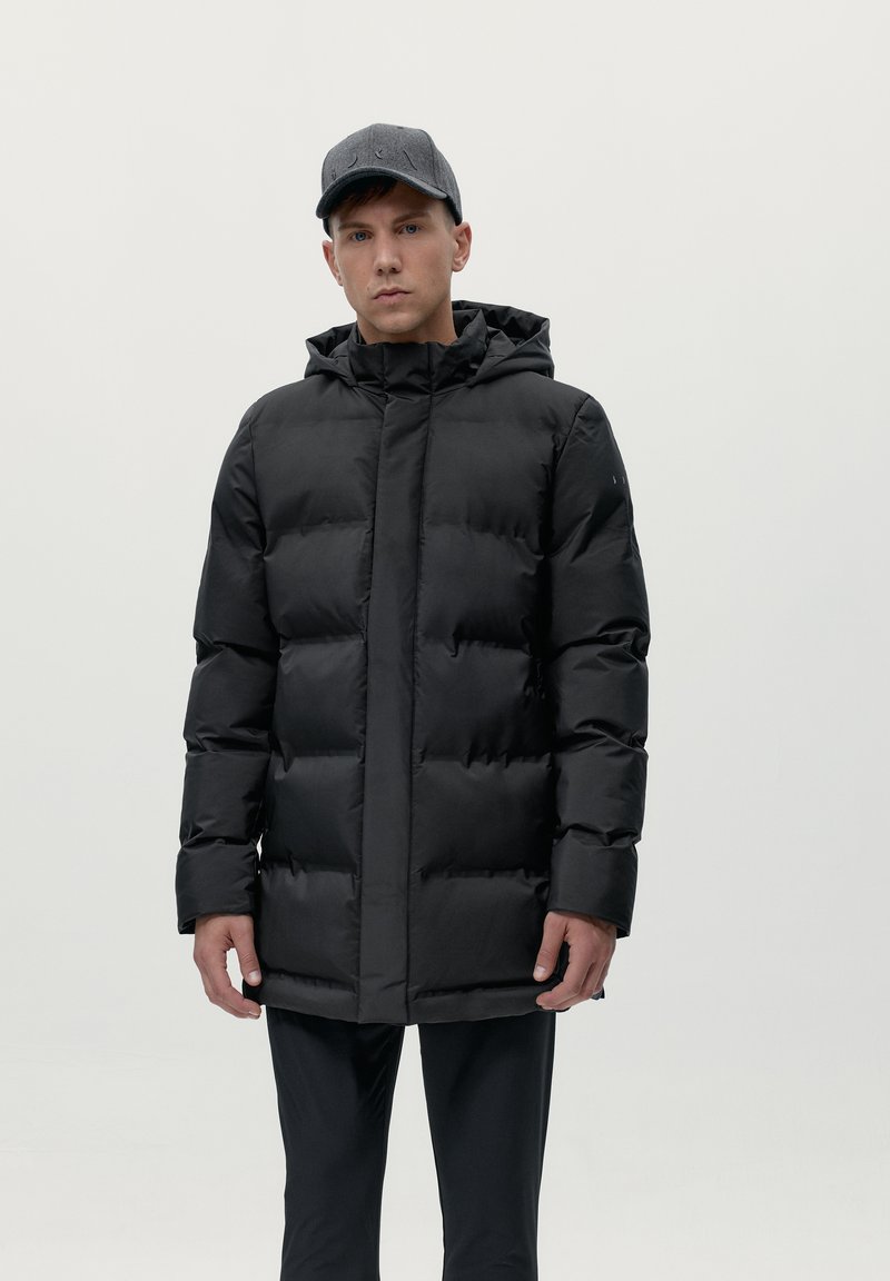 Melnā pūkainā jaka ar kapuci, ar kvadrātveida dizainu, gludu tekstūru un priekšējo rāvējslēdzēju. Kombinēta ar pelēku cepuri.