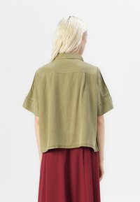 Korte mouwen, olijfgroen shirt van zachte stof, met een knoopsluiting bij de kraag en een ontspannen pasvorm, gecombineerd met een vloeiende bordeauxrode rok.