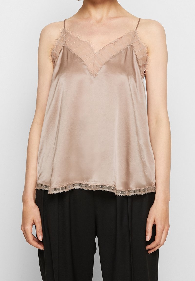 Femme portant un top en satin beige sans manches avec finition en dentelle et fines bretelles, associé à un pantalon noir sur un fond uni.