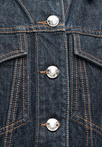 Σκούρο μπλε ύφασμα τζιν με τρία ασημένια μεταλλικά κουμπιά και πορτοκαλί ραφές γύρω από δύο θήκες στο στήθος ενός σακακιού.