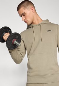 Khaki színű kapucnis pulóver kenguruzsebbel és húzózsinórral a kapucnin. Fekete "BORG" logóval rendelkezik, miközben fekete súlyzók emelésével foglalkozik.