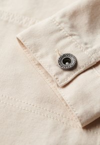 Beige bomuldsstof med synlige sømme og en металknap med et dekorativt præget bladdesign, placeret ved kraven.