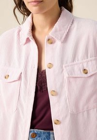 Lichtroze corduroy overhemdjack met een kraag, vier knopen en twee borstzakken. Draag het over een donkerpaarse cami.