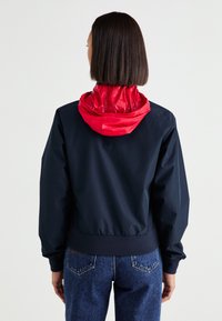 Chaqueta recortada azul marino con una capucha roja y brillante. Presenta puños y dobladillo elásticos, textura suave y una silueta casual.