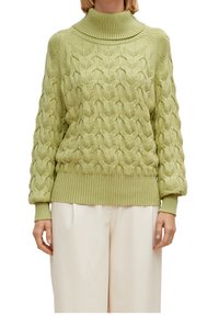 Grüner Kabelstrickpullover mit hohem geripptem Kragen und strukturiertem Muster. Gerippte Ärmelbündchen und Saum, kombiniert mit hellen Creme-Hosen.