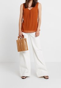 Blusa senza maniche marrone con dettagli a pieghe, abbinata a pantaloni bianchi larghi e una borsa in legno a righe. Sandali neri con cinturini completano il look.