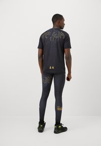 Schwarzes Sport-T-Shirt mit goldenen Designs und Schriftzügen auf dem Rücken, kombiniert mit schwarzen Leggings mit Akzenten und Grafiken, getragen mit schwarzen Schuhen.