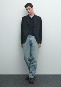 Schwarzer Blazer über einem dunklen V-Ausschnitt-Pullover, kombiniert mit hellblauen Jeans und braunen Schnürschuhen. Schlichte, klare Linien und weiche Texturen.