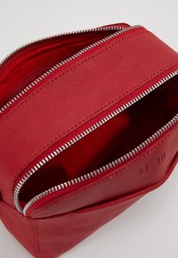 Punainen vetoketjullinen laukku, joka on valmistettu teksturoidusta materiaalista. Siinä on kaksi vetoketjua ja tilava sisäosa, jossa on kiinteä vuori.