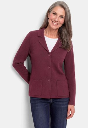 Vrouw met lang grijs haar, die een bordeauxrode cardigan met knopen draagt over een wit shirt en donkeblauwe spijkerbroek, glimlachend met één hand achter haar rug.