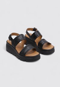 Sandalias de cuero negro con dos correas anchas, una hebilla ajustable y suela de plataforma. Forro interior marrón. Textura suave y diseño minimalista.