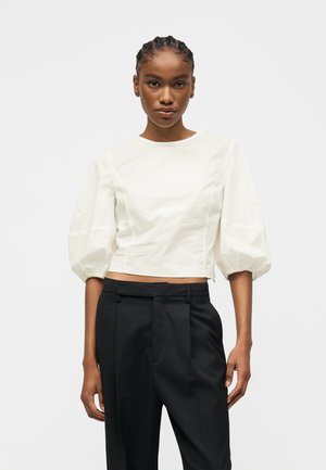 Femeie cu părul împletit, purtând un crop top alb cu mâneci bufante și pantaloni plisati negri cu talie înaltă, stând în fața unui fundal simplu.