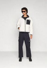 Krémfehér polárdzseki fekete mellzsebbel, fekete többzsebes cargo nadrággal és fekete sportcipővel párosítva.