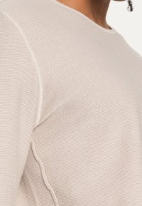 Hellbeiger Strickpullover mit strukturierter Oberfläche, mit rundem Halsausschnitt und sichtbaren Nähten an den Schultern.