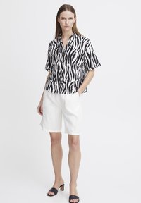 Camicia a maniche corte a righe bianche e nere con bottoni, abbinata a pantaloncini di lino bianchi e sandali neri con tacco. Sfondo semplice.