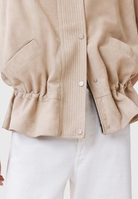 Gilet in suede beige con dettagli a coste, chiusure a scatto e orlo elasticizzato. Mostrato abbinato a pantaloni in denim bianchi.