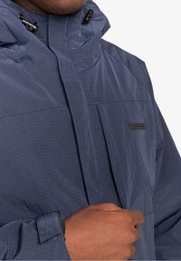 Granatowa kurtka w kratę, z wysokim kołnierzem, regulowanym kapturem i kieszenią na piersi z subtelnym logo. Lekki materiał.