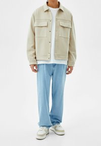 Beige fleece jacket with two chest pockets, button closure, and a relaxed fit. Paired with light blue jeans and white sneakers.