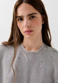 Sudadera gris con escote en V, adornada con piedras preciosas de colores dispersas en varias formas. Textura suave, diseño informal.