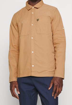 Mand iført en lys brun knap-up langærmet skjorte med forlommer og marineblå bukser mod en ensfarvet baggrund.