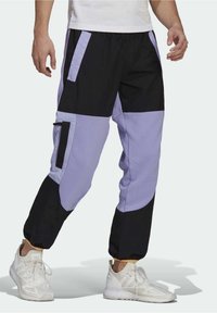 Man som bär svarta och lavendelfärgade färgblockerade joggerbyxor med cargo-fickor och vita sneakers, som står mot en enkel bakgrund.